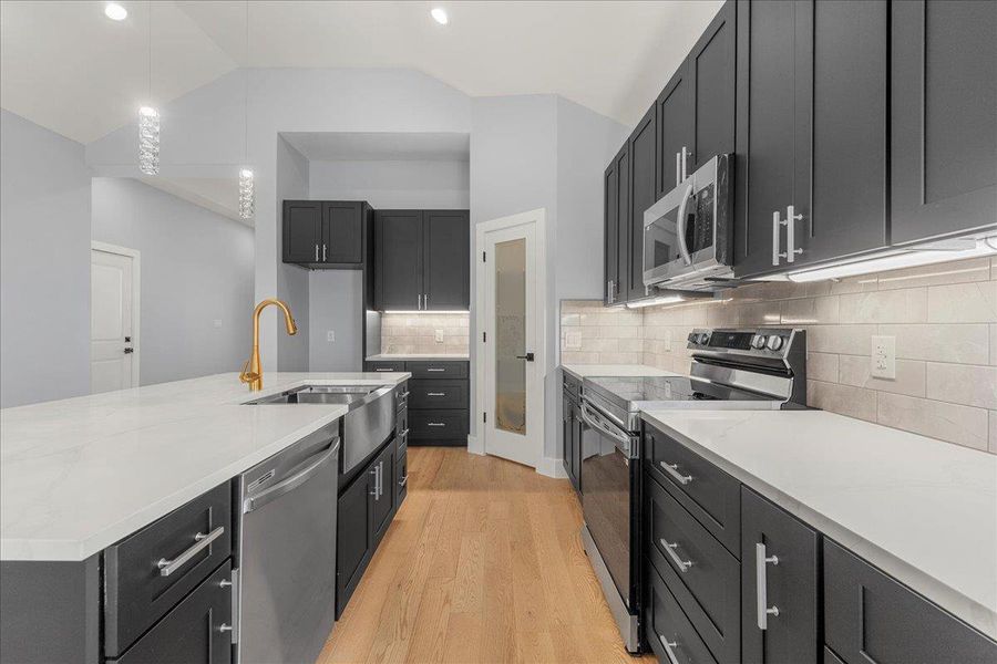 Kitchen with stainless steel appliances, dark cabinetry, a center island with sink, light stone counters, and vaulted ceiling Kitchen with stainless steel appliances, dark cabinetry, a center island with sink, light stone counters, and vaulted ceiling