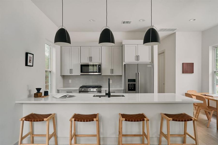 Modern kitchen featuring a large white countertop island with integrated sink, light gray cabinetry, stainless steel appliances, and sleek pendant lighting