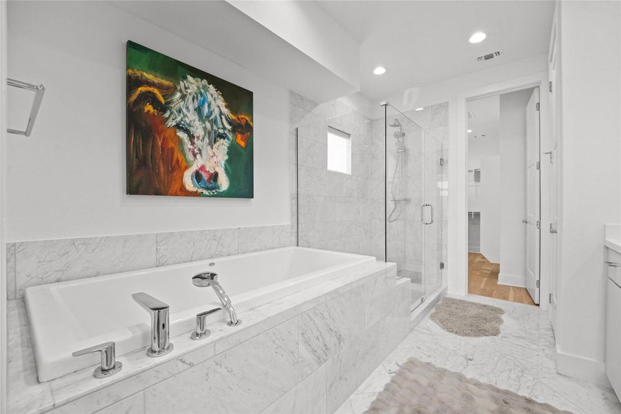 Recessed soaking tub with polished chrome fixtures, walk-in shower with frameless glass enclosure, marble-clad surfaces, and stone tile flooring