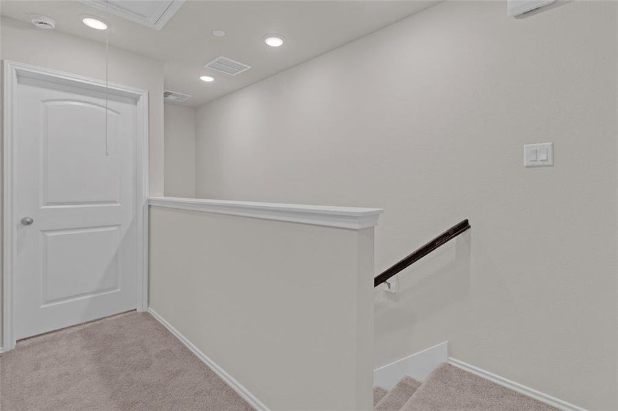 This photo shows a well-lit, neutral-toned stairway landing with beige carpeting, white trim, and a half-wall with a dark wooden handrail. Has modern recessed lighting in the ceiling.