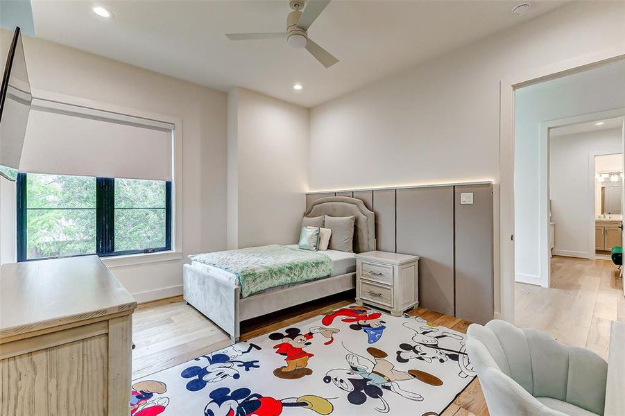 This is a cozy bedroom with modern design elements, featuring a stylish upholstered headboard with LED lighting, and a playful character-themed rug. The room is bright with natural light from a large window, has light wood flooring, and includes a ceiling fan for comfort.