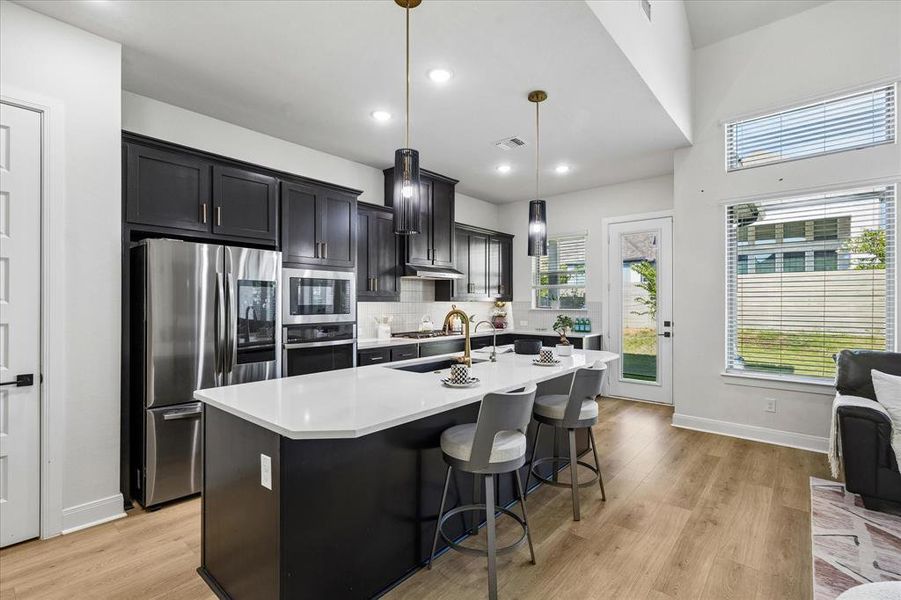 Open-concept kitchen featuring dark cabinetry, stainless steel appliances, a central island with a light-toned countertop, and gold-finish pendant lighting Open-concept kitchen featuring dark cabinetry, stainless steel appliances, a central island with a light-toned countertop, and gold-finish pendant lighting