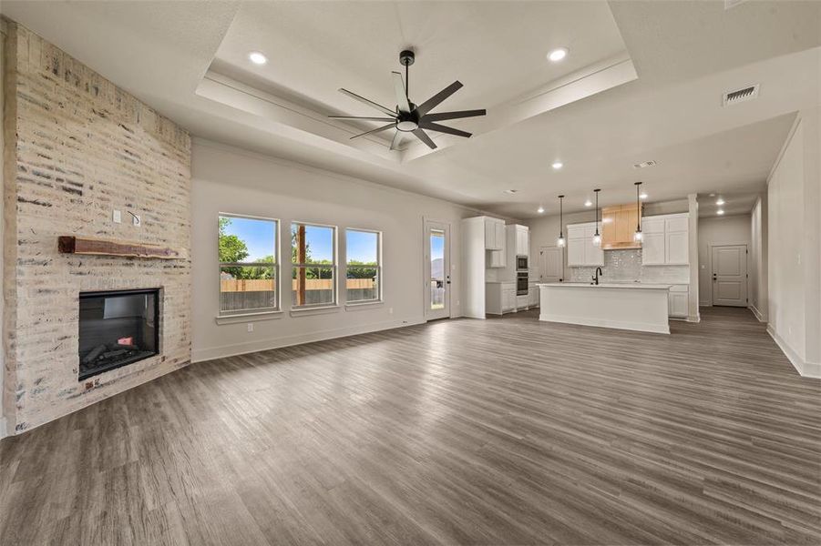Expansive interior featuring wood-finish flooring, a tray ceiling with a contemporary fan, and a painted brick fireplace with a wood mantel Expansive interior featuring wood-finish flooring, a tray ceiling with a contemporary fan, and a painted brick fireplace with a wood mantel