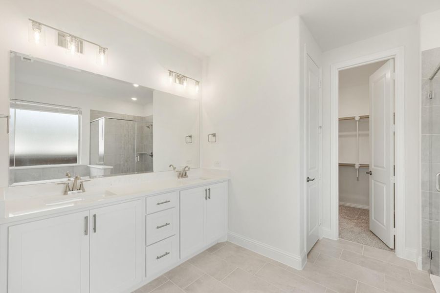Master bathroom with double sink vanity, white cabinets, large mirror, and walk-in closet entrance visible through open door.