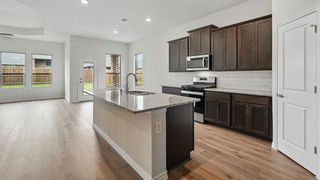 Open-concept kitchen featuring an island with a built-in sink, granite countertops, dark wood cabinetry, stainless steel appliances, and wood-finish flooring