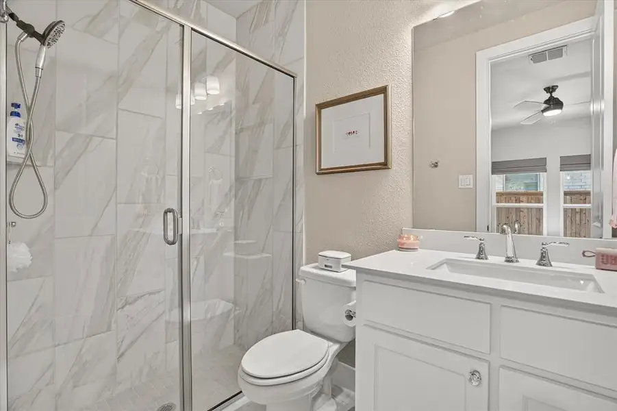 Bathroom featuring a glass-enclosed shower with white and gray tiled walls, a white vanity with an integrated sink and chrome fixtures, a large wall-mounted mirror, and a white toilet Bathroom featuring a glass-enclosed shower with white and gray tiled walls, a white vanity with an integrated sink and chrome fixtures, a large wall-mounted mirror, and a white toilet