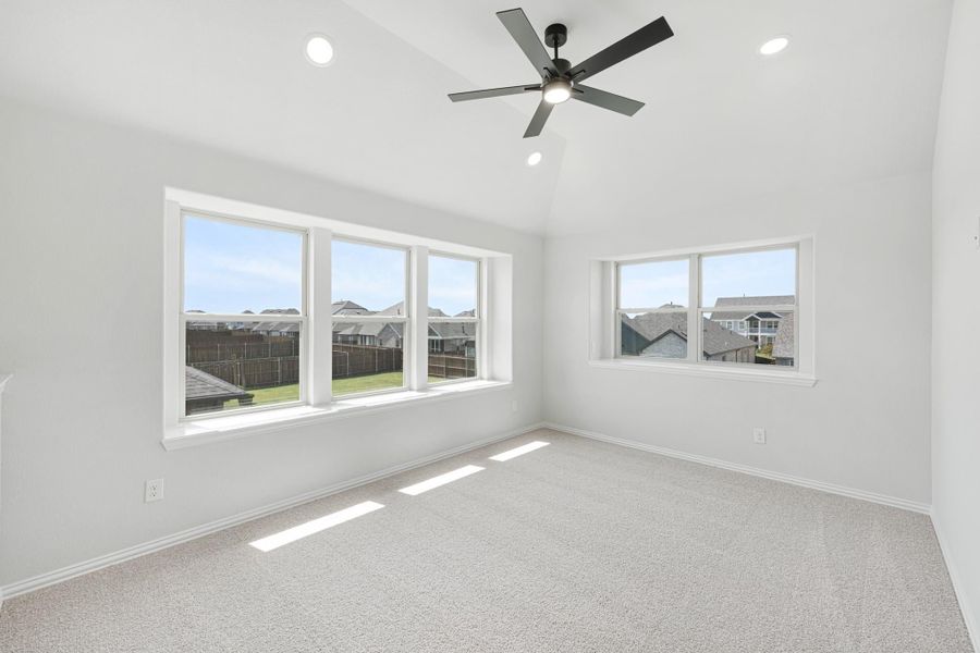 Empty bedroom with vaulted ceiling, ceiling fan, carpet flooring, and large windows overlooking a neighborhood Empty bedroom with vaulted ceiling, ceiling fan, carpet flooring, and large windows overlooking a neighborhood