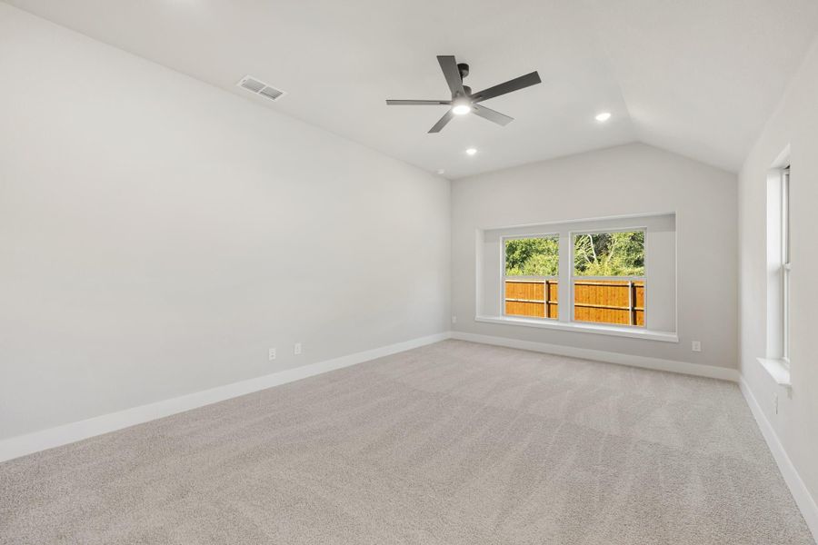 Empty bedroom with vaulted ceiling, ceiling fan, carpet flooring, and a window overlooking a backyard fence.