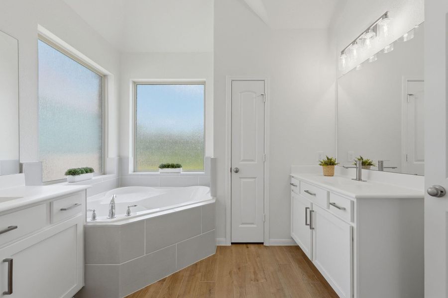 This bathroom features a spacious corner bathtub with large frosted windows, dual sinks with ample counter space, sleek white cabinetry, modern fixtures, and a warm wood tile floor.