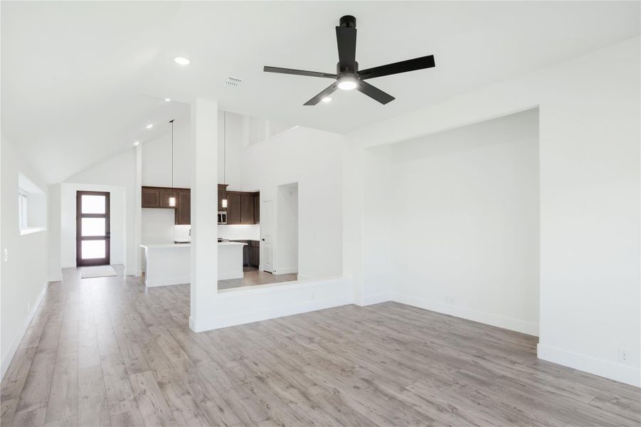 Open living area with vaulted ceiling, black ceiling fan, wood floors, and kitchen visible in background Open living area with vaulted ceiling, black ceiling fan, wood floors, and kitchen visible in background