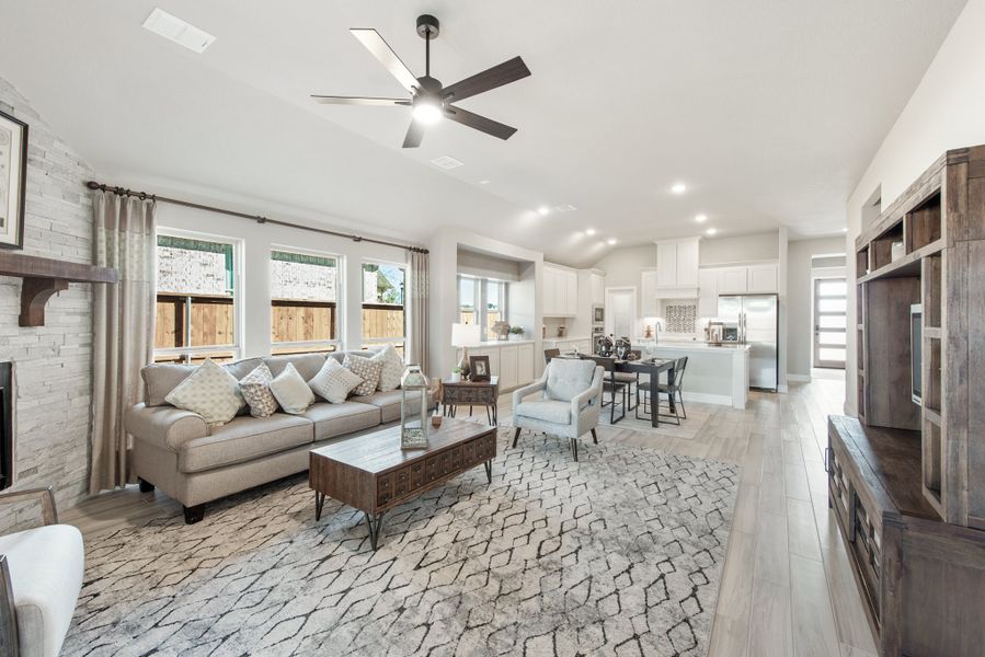 Open-concept living room with stone fireplace, ceiling fan, and hardwood floors flowing into kitchen