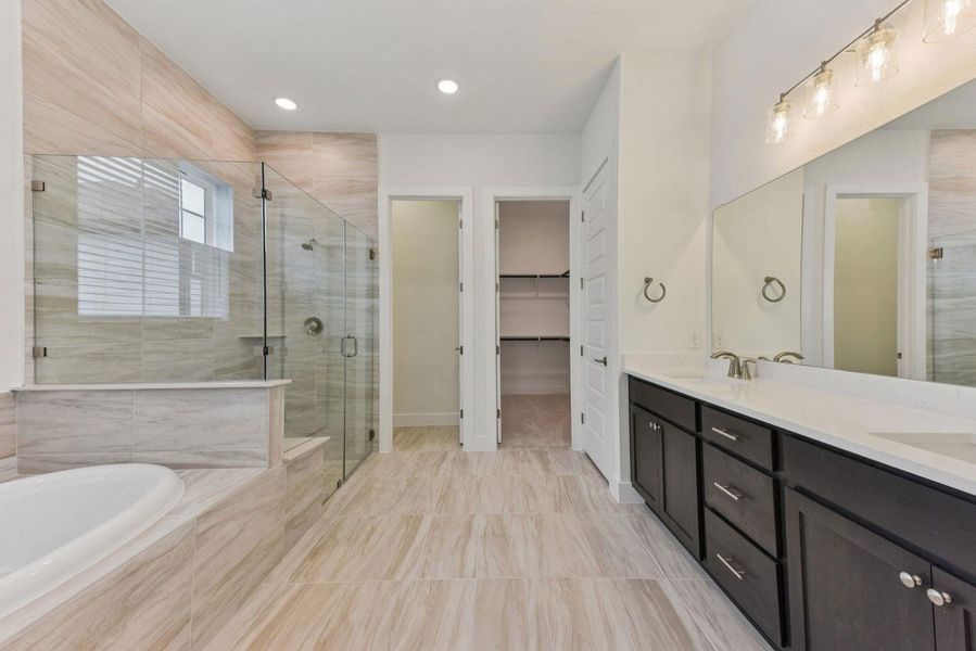Spacious bathroom featuring a dual vanity with dark wood cabinetry and white countertops, a large mirror with contemporary lighting, a glass-enclosed shower with a built-in bench, a separate soaking tub, and neutral-toned tiled flooring and shower surround
