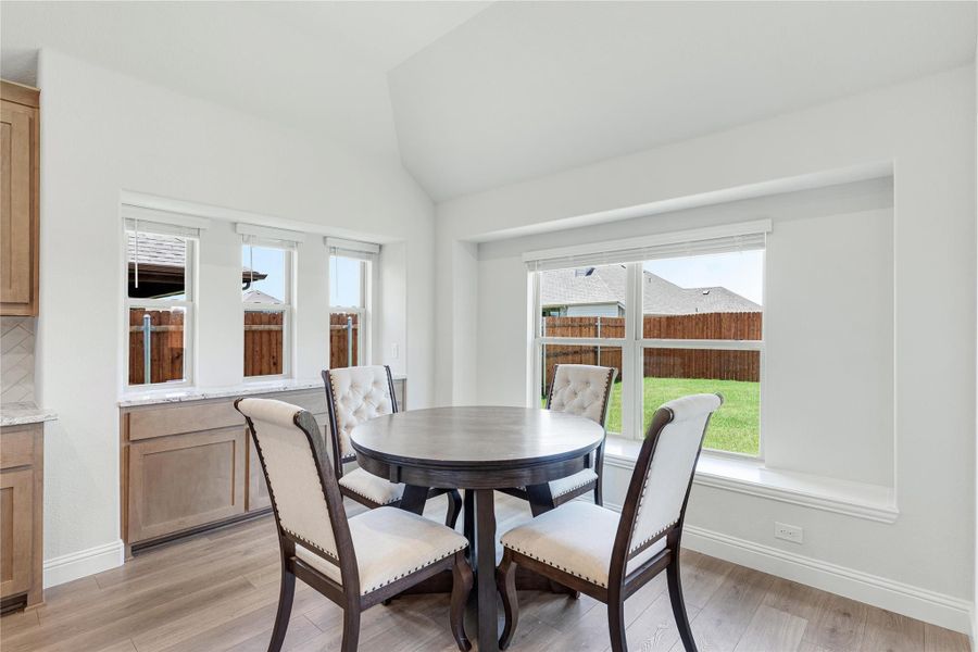 Breakfast nook with round dark wood table, four upholstered chairs, vaulted ceiling, and large windows overlooking backyard.