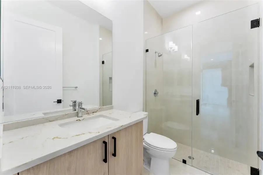 Bathroom featuring a vanity with light wood-finish cabinetry, a stone-surface countertop with an integrated rectangular sink, a large wall-mounted mirror, and a glass-enclosed shower with a recessed wall niche