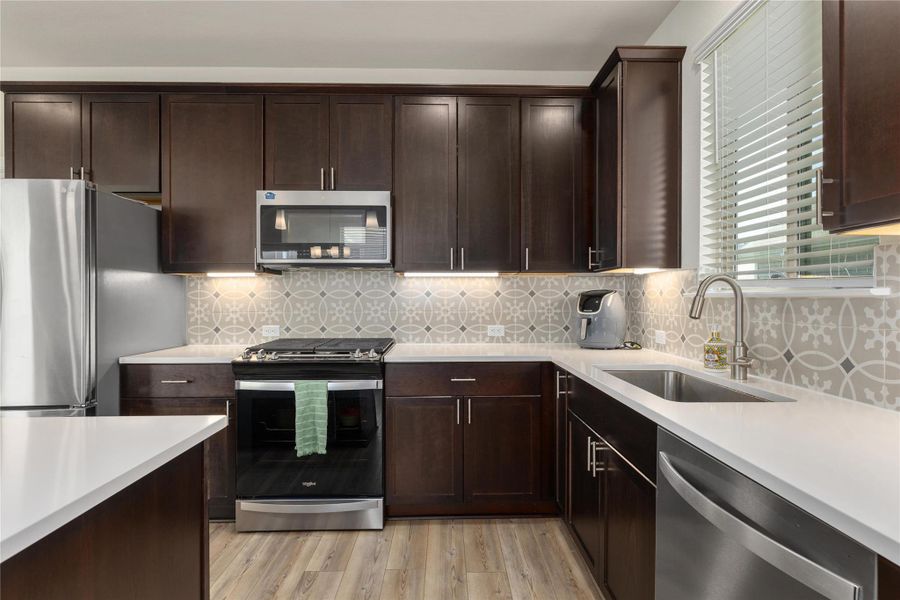 Kitchen featuring appliances with stainless steel finishes, backsplash, dark brown cabinetry, light wood finished floors, and light stone countertops