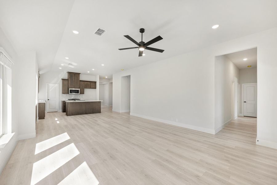 Open living area with light wood floors, ceiling fan, and kitchen with dark cabinets and island in background