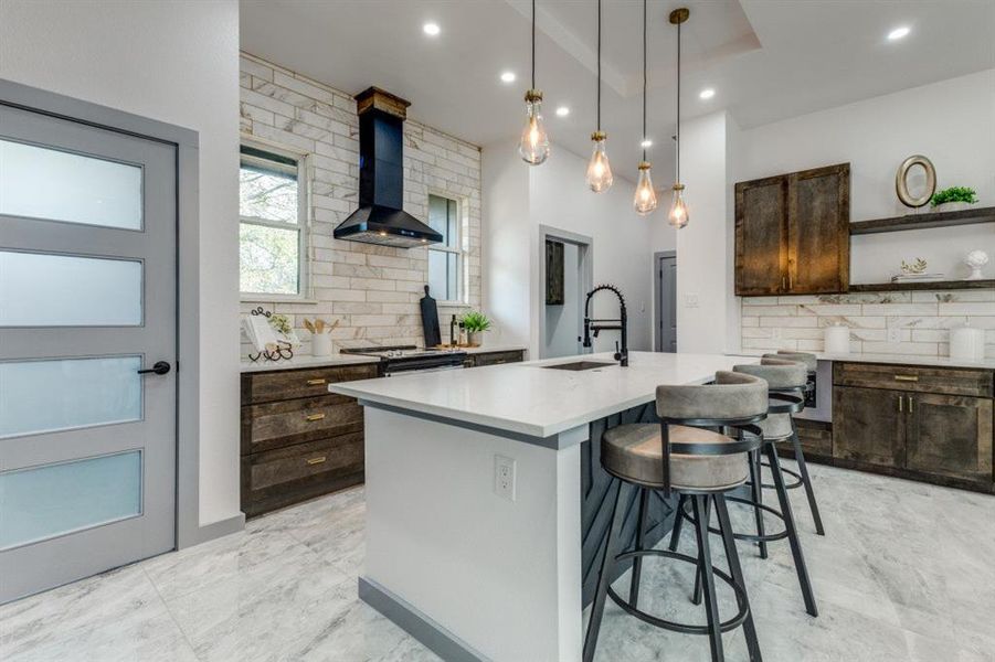 Kitchen with sink, tasteful backsplash, an island with sink, wall chimney range hood, and pendant lighting