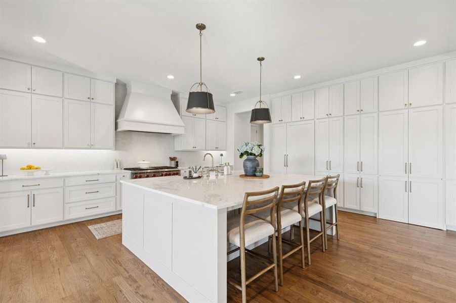 Kitchen featuring a breakfast bar, a kitchen island with sink, custom exhaust hood, hanging light fixtures, and light wood-style flooring Kitchen featuring a breakfast bar, a kitchen island with sink, custom exhaust hood, hanging light fixtures, and light wood-style flooring