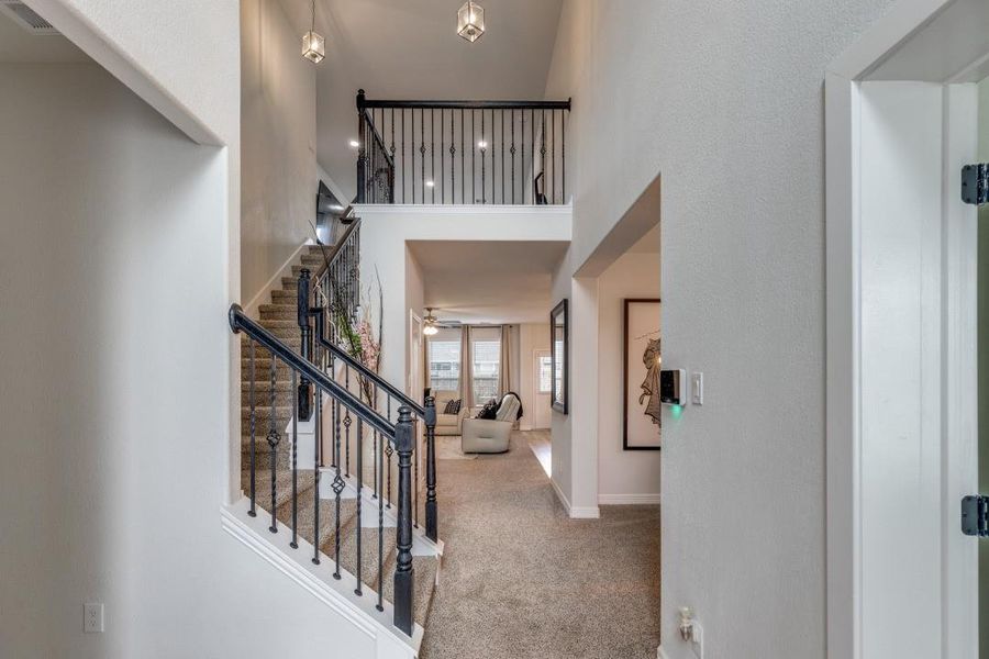 A modern, open-concept home interior with a welcoming entryway. It features a staircase with elegant iron railings leading to an upper level and a view into a cozy living area with carpeted floors and abundant natural light.