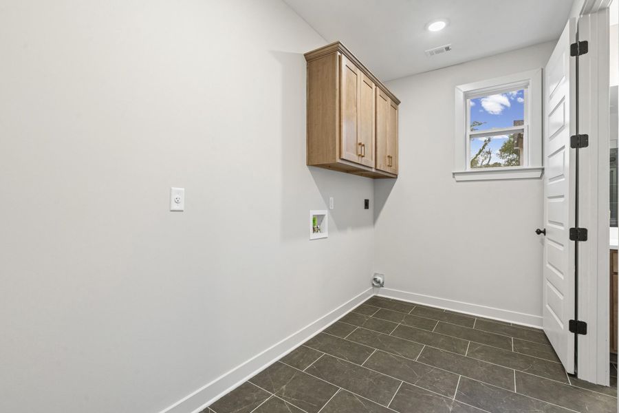 Utility room featuring dark rectangular tile flooring, light wood-finish cabinetry, a white-framed window, and a white paneled door with matte black hardware Utility room featuring dark rectangular tile flooring, light wood-finish cabinetry, a white-framed window, and a white paneled door with matte black hardware