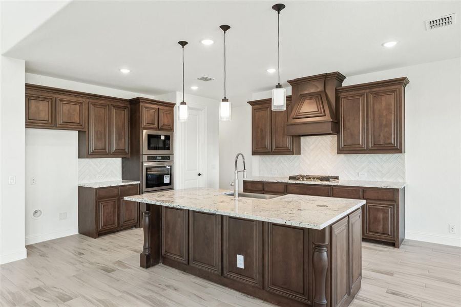 Kitchen with dark wood cabinets, large granite island, pendant lights, and stainless steel appliances on light hardwood floors Kitchen with dark wood cabinets, large granite island, pendant lights, and stainless steel appliances on light hardwood floors