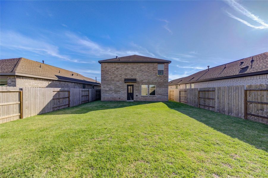 This photo showcases a spacious backyard with a well-maintained lawn, bordered by a wooden fence for privacy. The brick exterior of the two-story home is visible, offering a clear view of the house's rear and neighboring homes. Ideal for outdoor activities and relaxation.