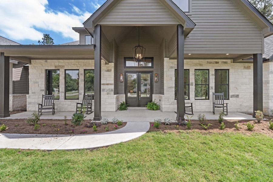 Impressive front elevation with a covered entry framed by stone and rich wood accents. Double front doors with glass panels are highlighted by statement lighting and bold columns, creating a strong architectural presence. Symmetrical windows and clean landscaping complete the look, delivering standout curb appeal from the moment you arrive.