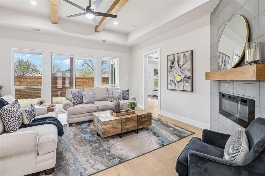 Open-concept living area featuring a tray ceiling with wood beams, light wood-finish flooring, and a modern fireplace with a tiled surround and wood mantel