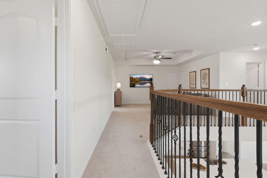 This photo showcases a bright, carpeted upstairs hallway with elegant iron and wood railing overlooking a lower level. The space is well-lit with recessed lighting and a ceiling fan, creating a welcoming and airy atmosphere.