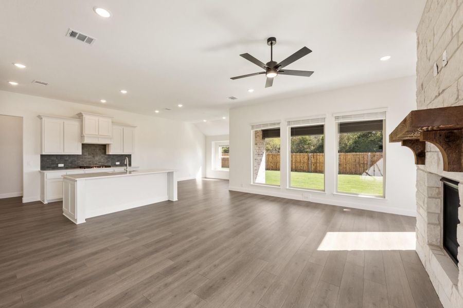 Open-concept living area with stone fireplace, ceiling fan, hardwood floors, and kitchen with white cabinets and island Open-concept living area with stone fireplace, ceiling fan, hardwood floors, and kitchen with white cabinets and island
