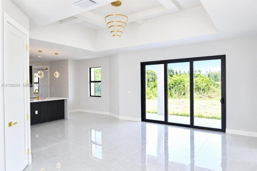Tray ceiling with multi-tiered gold-tone chandelier, large format polished tile flooring, sliding glass doors with black frames, and kitchen island featuring a black fluted facade