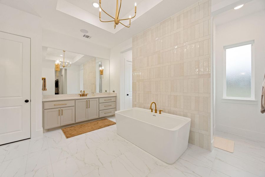 This primary bathroom features a modern design with a freestanding bathtub, elegant gold fixtures, a large vanity, and a spacious mirror. The room is bright and airy, with neutral tones and stylish tile work.