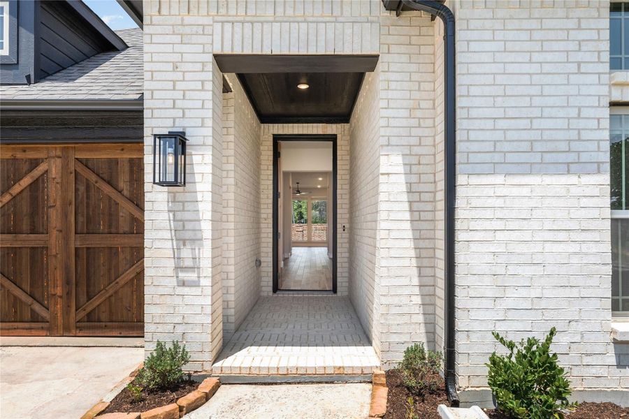 Front entry of white brick home with black-framed door, wood ceiling alcove, and wall lantern beside wood garage door