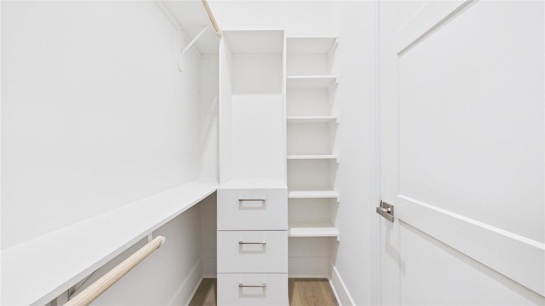 This photo shows a bright, modern walk-in closet with ample shelving, hanging space, and three drawers, all in a sleek white finish.
