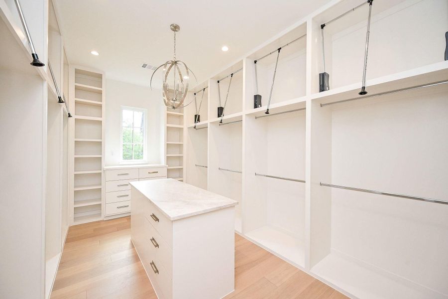 Primary Walk-in closet-spacious and Luxurious with Over-Under Custom Cabinetry. Island and Chandelier Accent. featuring open shelves, hanging rods, and a central island with drawers.
