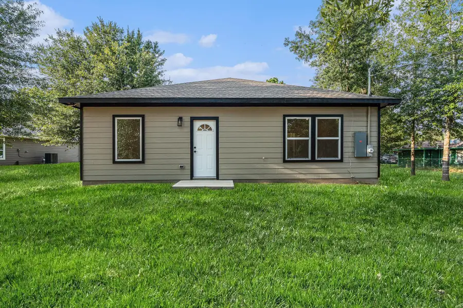This photo shows a charming, single-story home with a simple, modern exterior. It features a dark roof, neutral siding, and contrasting window frames. The house is surrounded by a lush, green lawn and is set against a backdrop of trees, offering a peaceful and private setting.