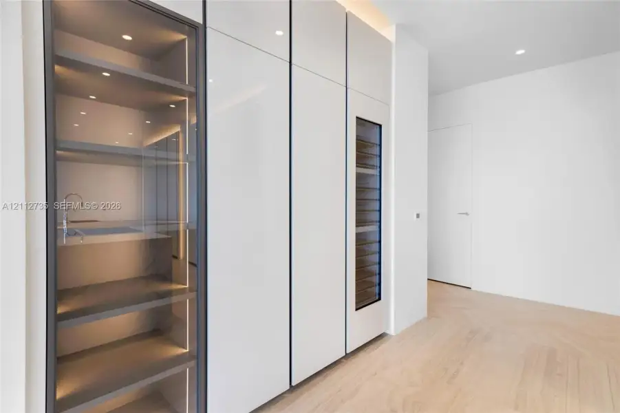 Custom millwork featuring floor-to-ceiling high-gloss cabinetry, integrated wine refrigeration, glass-front display shelving with LED illumination, and chevron wood-finish flooring