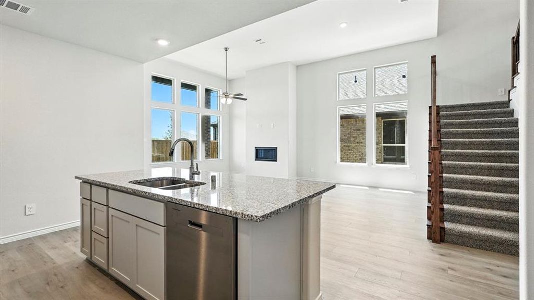 Spacious open-concept living area featuring an island kitchen with granite countertops and stainless steel appliances, wood-finish flooring, a contemporary fireplace, and a carpeted staircase with wood railing