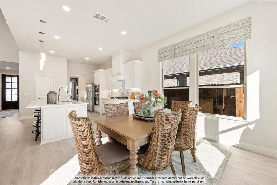 Open dining area with wood table and wicker chairs, adjacent to white kitchen with island and stainless appliances