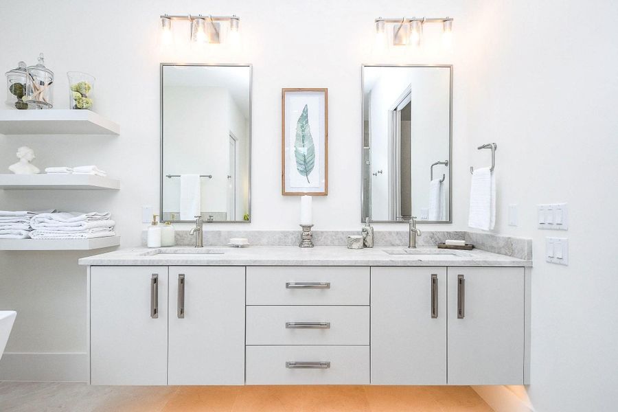 This bathroom features a modern double vanity with sleek gray countertops, two large mirrors, and ample storage. Bright lighting and minimalist shelves add to the clean, contemporary design.