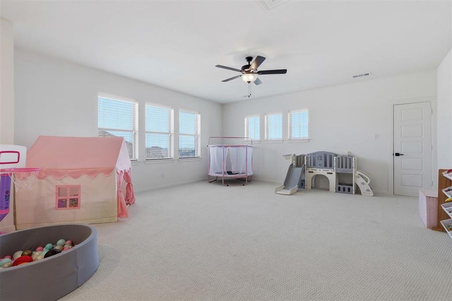 Spacious room with light-colored carpeting, multiple windows with blinds, a ceiling fan with integrated lighting, and a white paneled door with matte black hardware Spacious room with light-colored carpeting, multiple windows with blinds, a ceiling fan with integrated lighting, and a white paneled door with matte black hardware