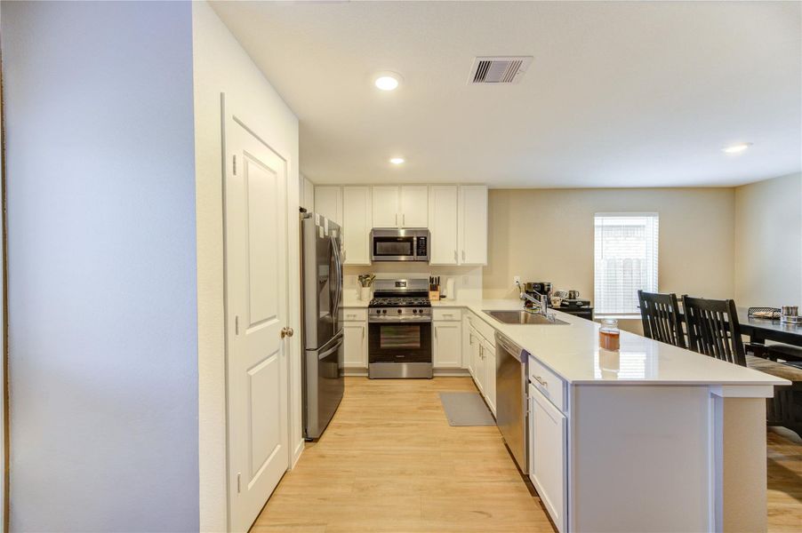 This kitchen features modern stainless steel appliances, white cabinetry, and a spacious island with a sink. The open layout flows into a cozy dining area with natural light from a large window.