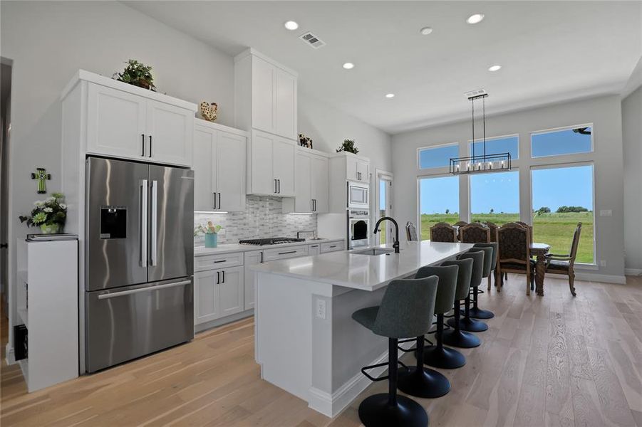 Kitchen with stainless steel appliances, light wood-style flooring, a chandelier, an island with sink, and decorative backsplash Kitchen with stainless steel appliances, light wood-style flooring, a chandelier, an island with sink, and decorative backsplash