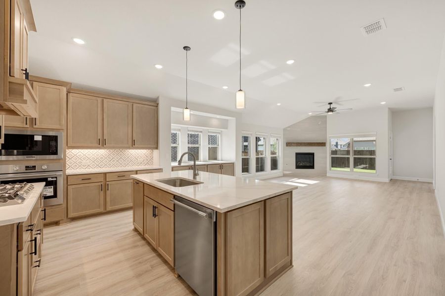 Open-concept kitchen with light wood cabinets, center island, and stainless appliances opening to living room with fireplace