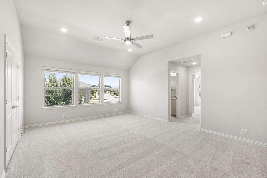 Spacious bedroom with vaulted ceiling, ceiling fan, carpet flooring, and three windows overlooking trees