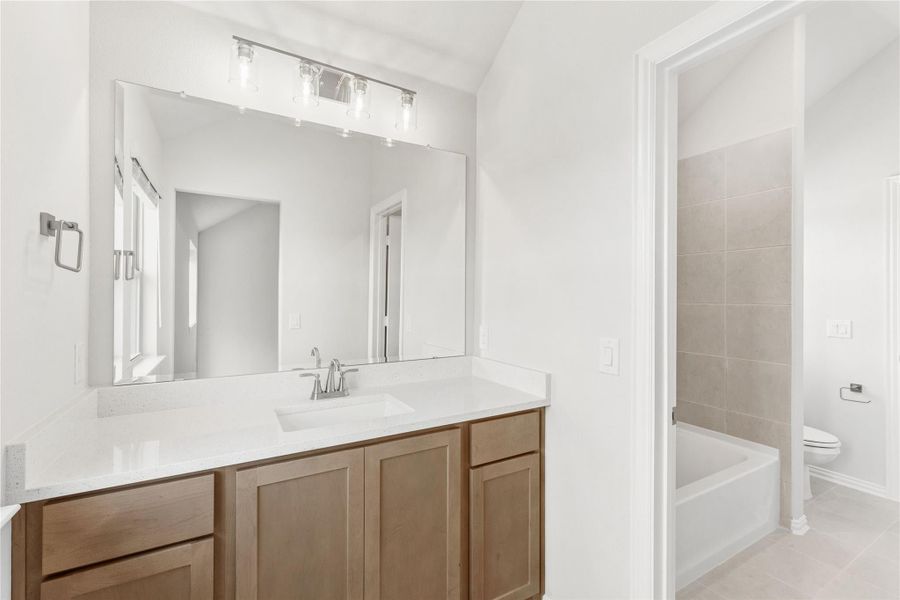 Bathroom with wood vanity, white quartz countertop, large mirror, and tiled tub-shower combo visible through doorway.