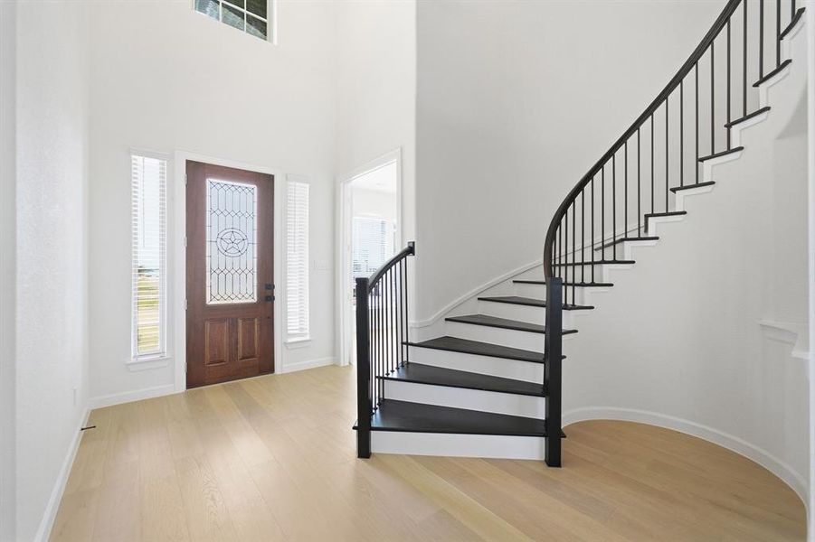 Grand foyer featuring a curved staircase with black metal balusters, a decorative wood-panel entry door with leaded glass, tall windows, and light wood-finish flooring