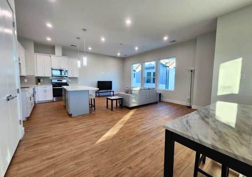 This photo showcases a modern, open-concept living space with a kitchen and living room. The kitchen features white cabinets, stainless steel appliances, and an island with pendant lighting. The living room includes large windows for natural light and wood flooring throughout. This photo showcases a modern, open-concept living space with a kitchen and living room. The kitchen features white cabinets, stainless steel appliances, and an island with pendant lighting. The living room includes large windows for natural light and wood flooring throughout.