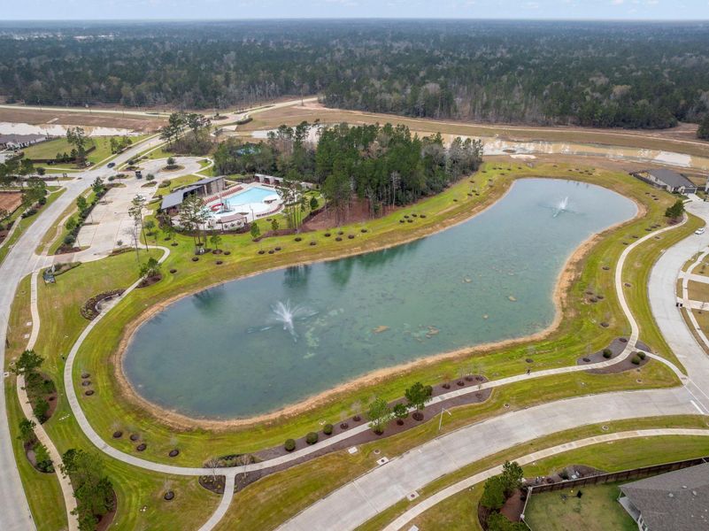 This aerial view showcases a serene residential neighborhood with a large pond featuring fountains, surrounded by lush greenery. A community center with a swimming pool is nestled among trees, offering recreational amenities. The area is well-planned with wide streets and landscaped pathways, providing a peaceful and inviting environment.