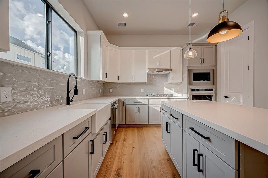 This modern kitchen features sleek white cabinetry, a stylish tile backsplash, and stainless-steel appliances. It has ample countertop space, pendant lighting, and a large window for natural light. The wood flooring adds warmth and elegance to the space.